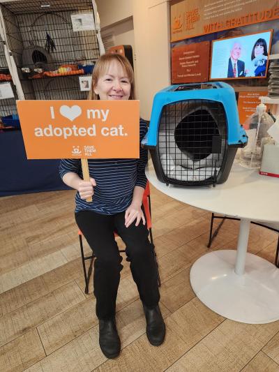 Smokey the cat in a carrier with his adopter sitting next to him holding a sign that says, I heart my adopted cat